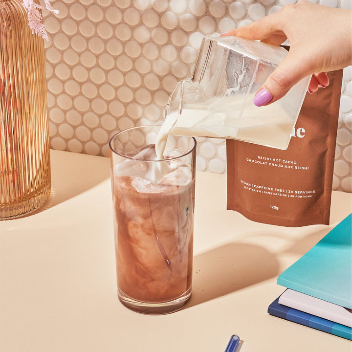 A hand pours milk into a glass of iced chocolate drink next to a packet and a vase.